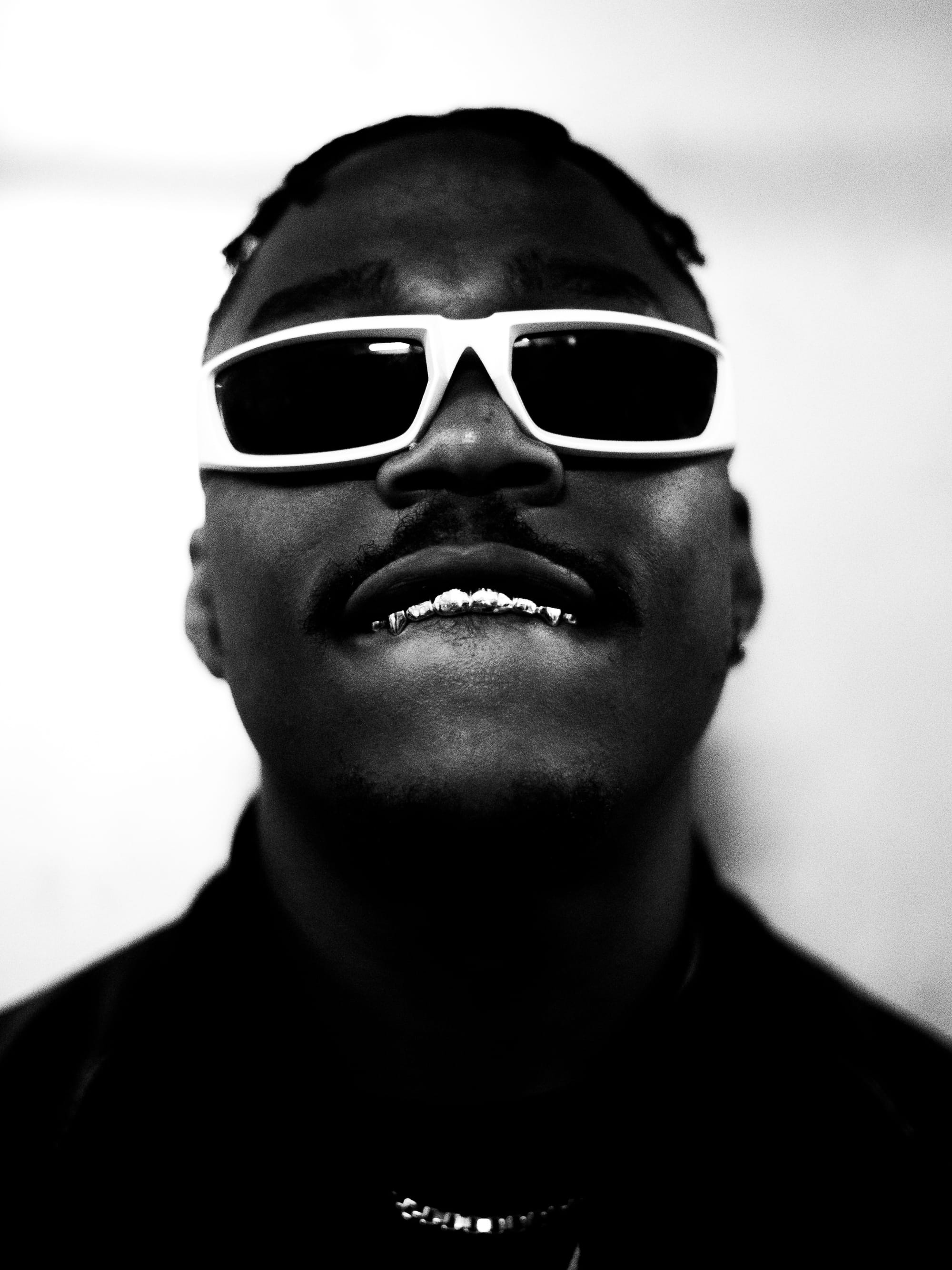 High-contrast black-and-white close-up portrait of a man wearing white sunglasses and grills, looking directly at the camera.