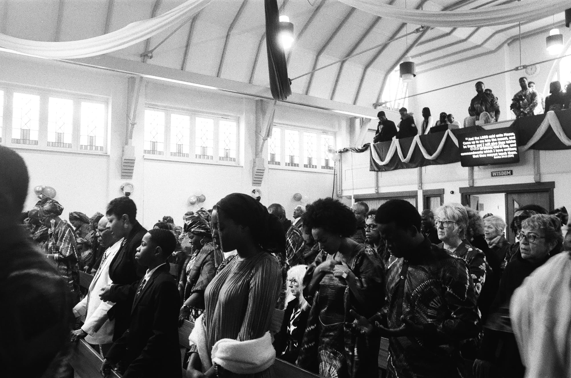 Black-and-white image of a church congregation standing and praying during a service, with people filling both the ground floor and balcony.