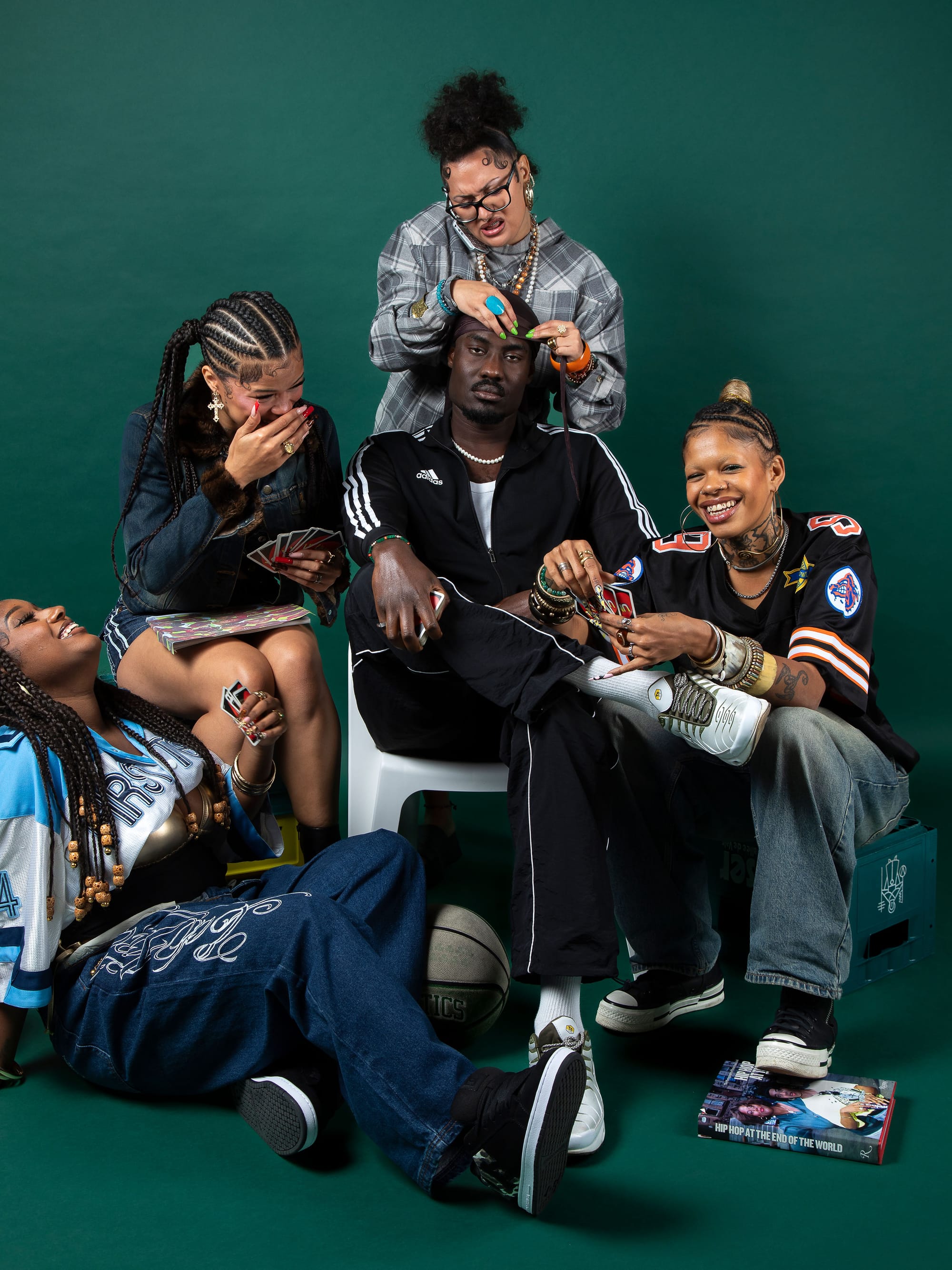 A group of five people posing playfully in front of a green backdrop, braiding hair, laughing, and playing cards in a stylised portrait.