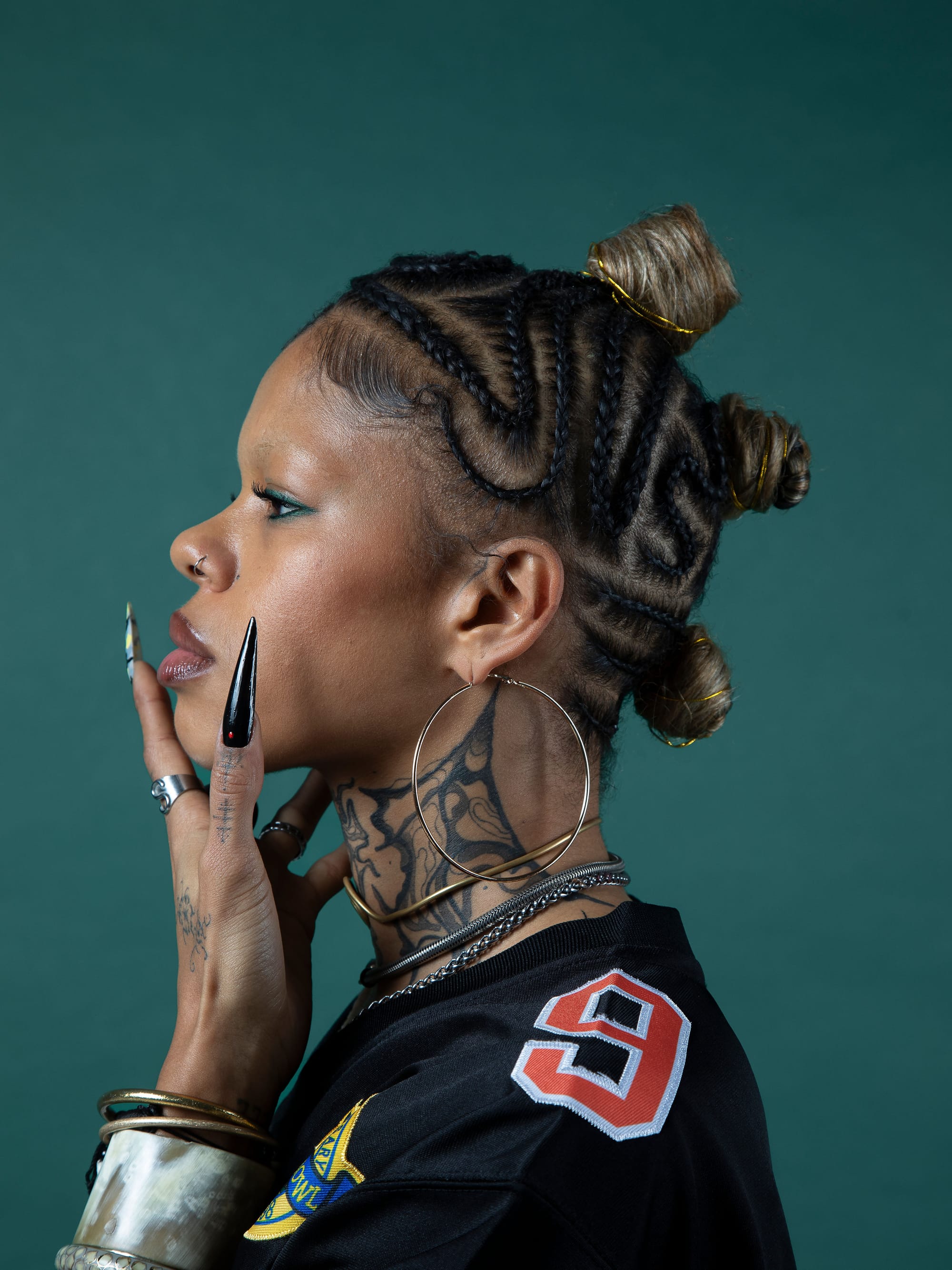 Profile portrait of a woman with intricate braided hair, large hoop earrings, and detailed tattoos, posed against a dark green backdrop.