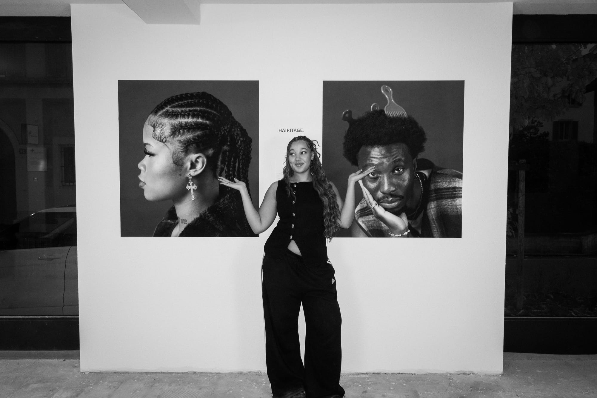 Black-and-white photo of an artist standing in a gallery between two large portraits from her “Hairitage” series.