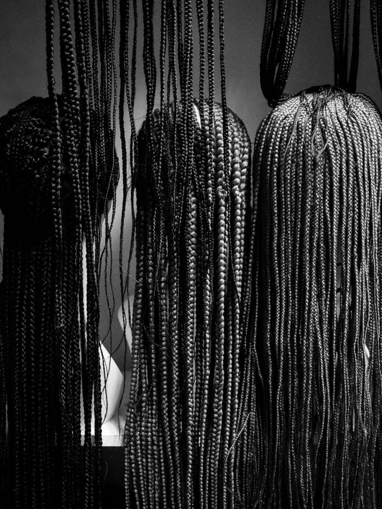 Black-and-white photograph of intricately braided hairstyles hanging forward, obscuring the faces behind them.